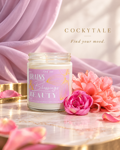 A glass jar candle labeled 'COCKYTALE BRAINS & Blessings BEAUTY' sits on a marble tray with pink flowers and draped lavender fabric in the background. The brand name 'COCKYTALE' and tagline 'Find your mood.