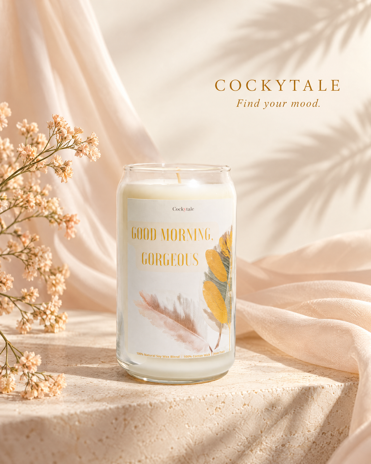 A glass jar candle labeled 'COCKYTALE' and 'GOOD MORNING GORGEOUS' sits on a textured beige surface with draped cream fabric and soft leaf shadows in the background. Small dried flowers appear at the left edge.