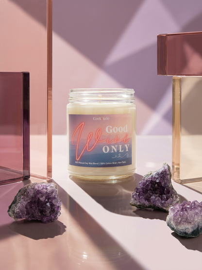 A jar candle labeled 'Good Wine ONLY' sits on a reflective surface between geometric glass blocks and purple crystal clusters, with soft pink and lavender lighting.