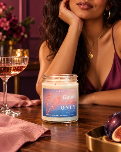 A jar candle labeled 'Good wine ONLY' sits on a wooden table in a cozy, dimly lit room with a woman in a purple top resting her head on her hand nearby. A filled wine glass, pink napkin, and figs are also visible.